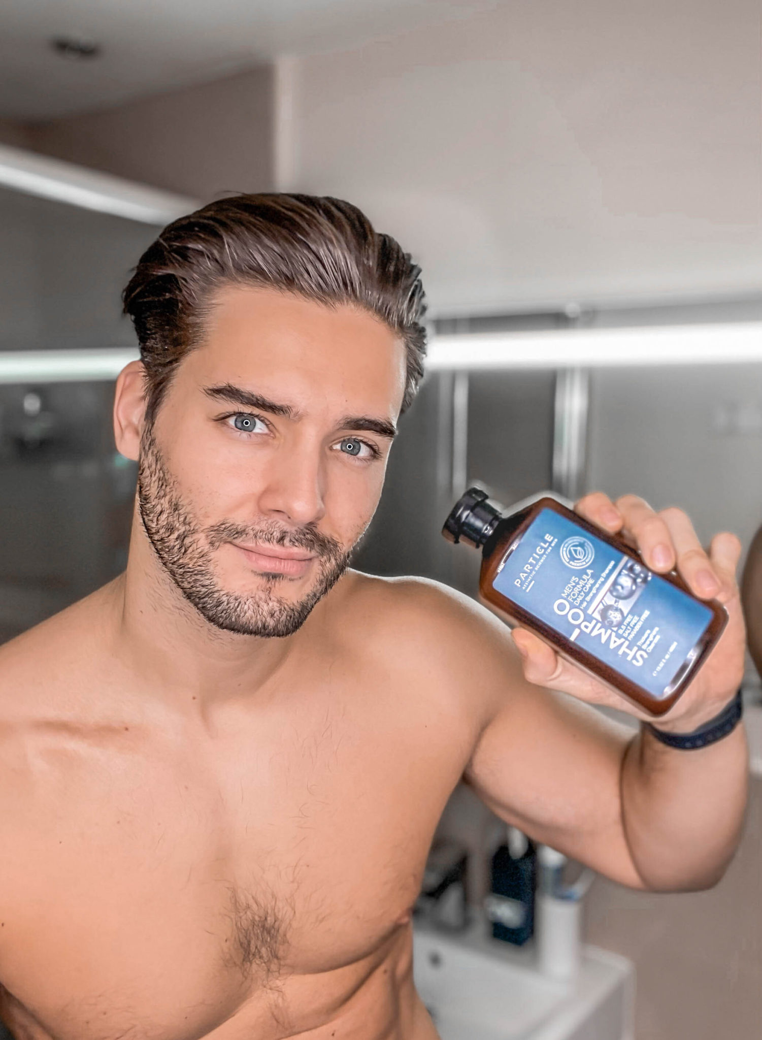 Man holding Particle Daily Care shampoo bottle, shirtless in a modern bathroom.