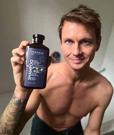 Smiling man with tattoo holding a Particle shampoo bottle.