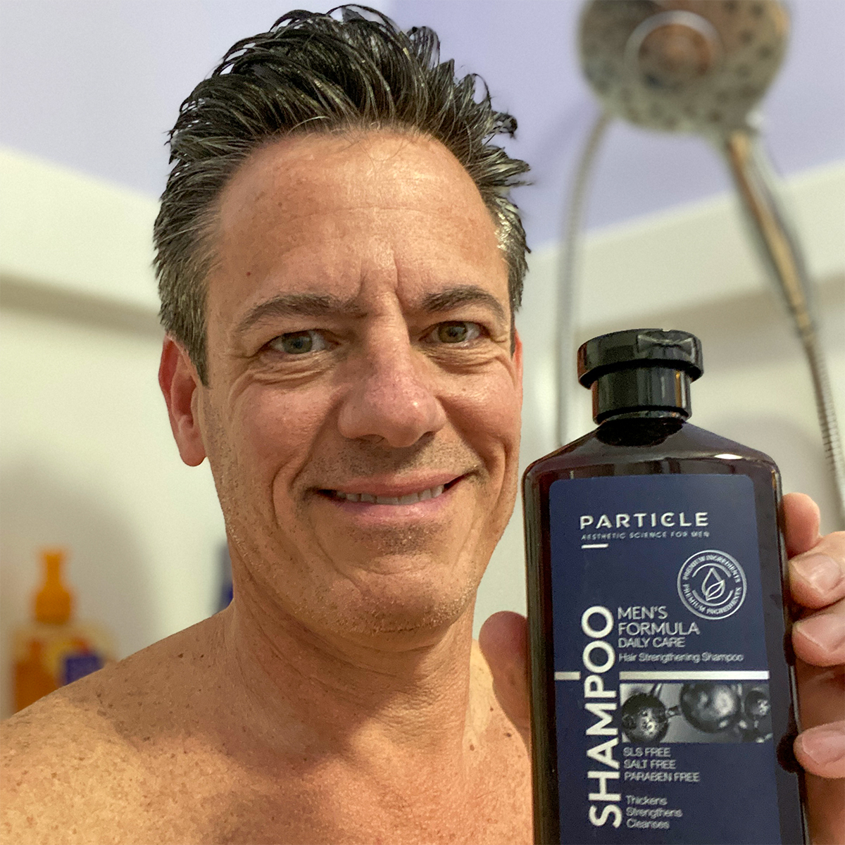 A shirtless man smiling, holding a brown bottle of Particle Men's Formula Shampoo with blue label.