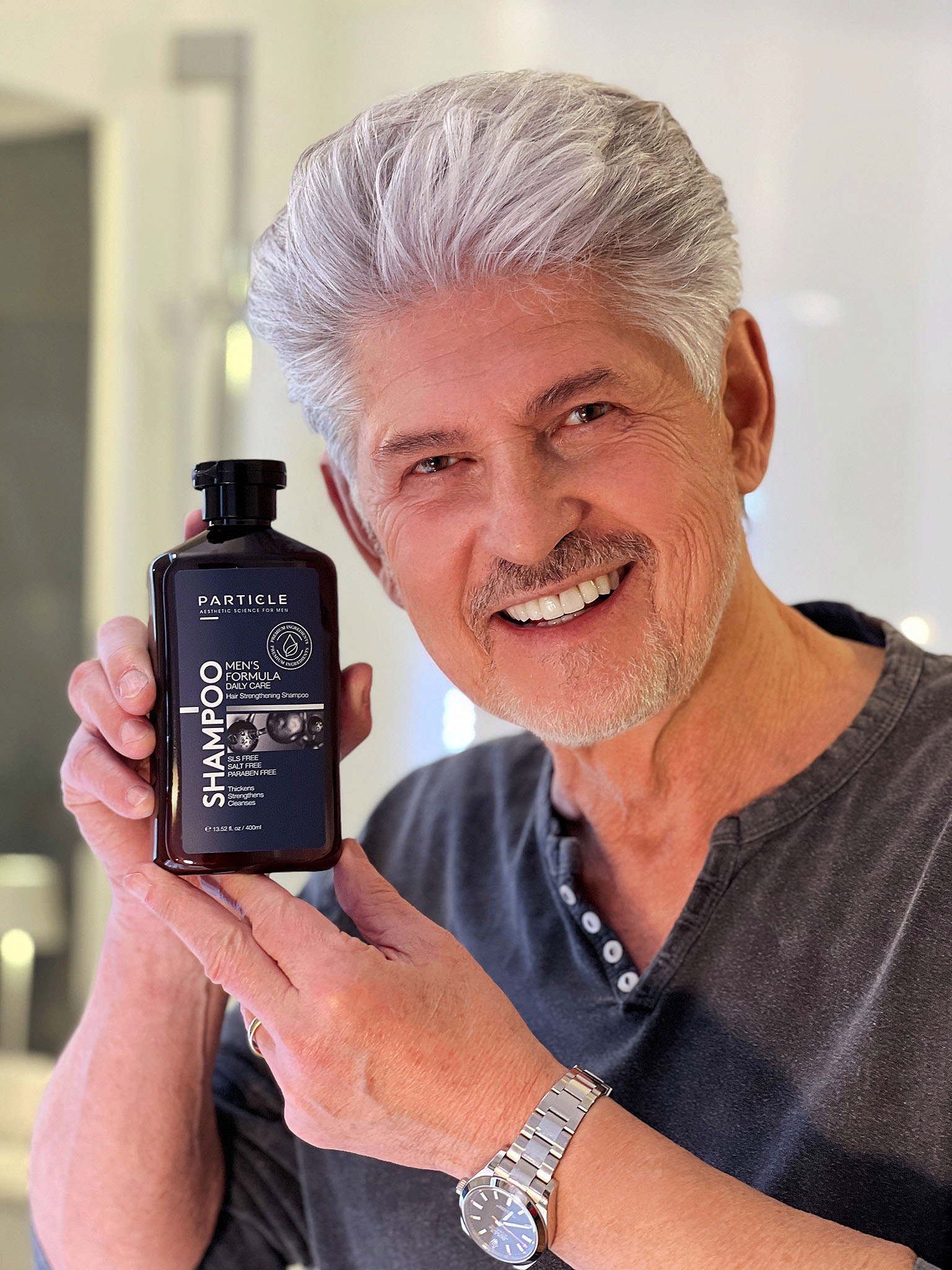 Man smiling and holding Particle Men's Formula Daily Care Strengthening Shampoo bottle.