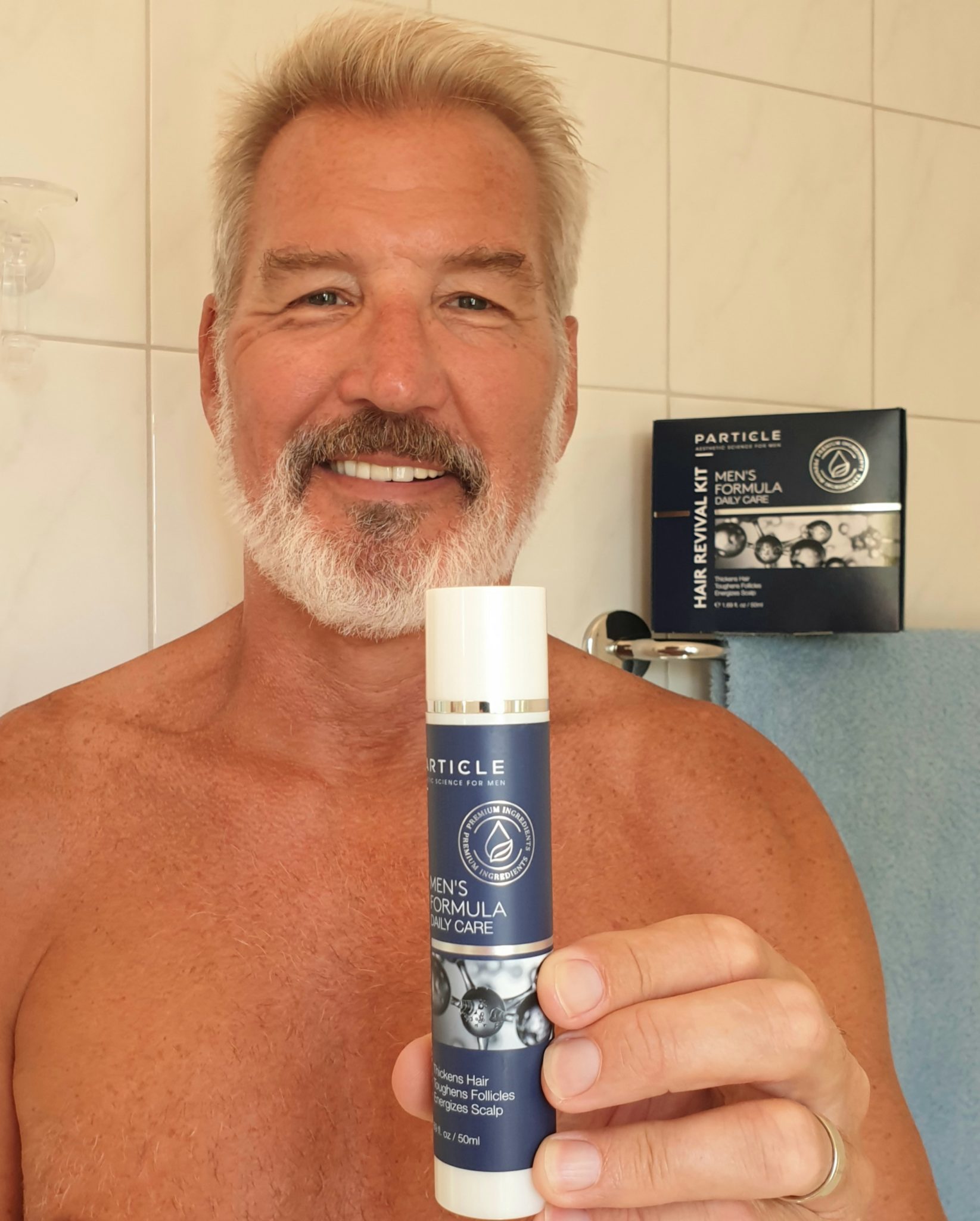 Man smiling, holding Particle Men's Formula Daily Care bottle in bathroom.