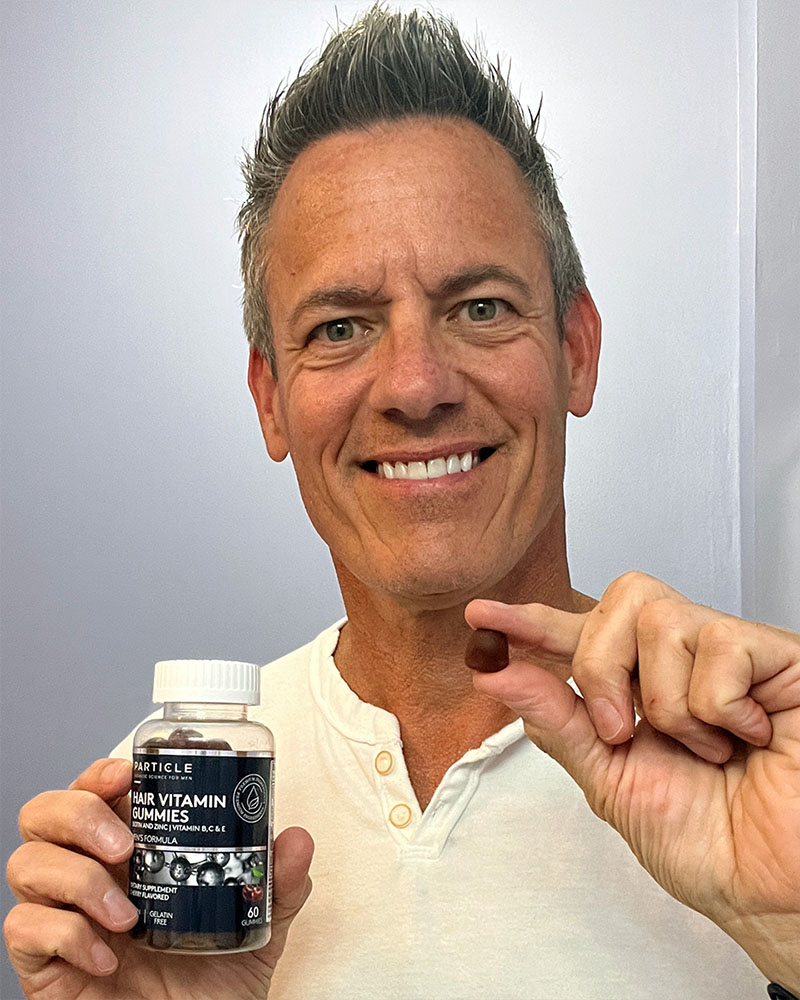 A man holds a gummy and a bottle of Particle Hair Vitamin Gummies with a smile.