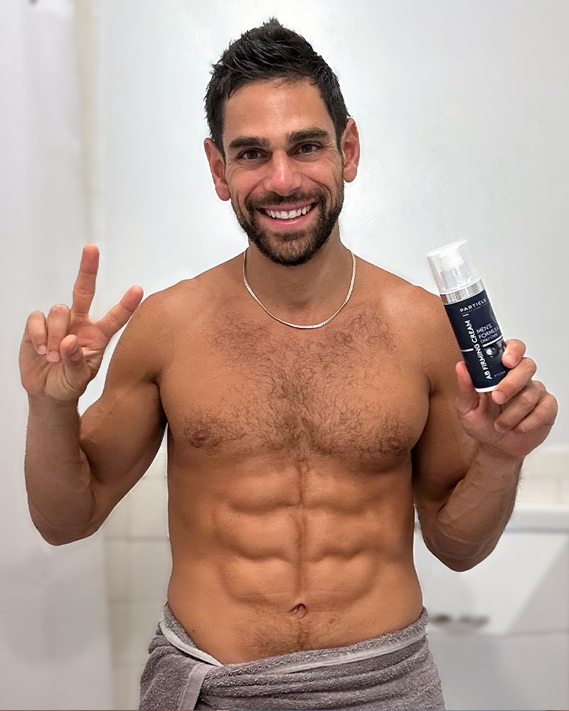 Shirtless man holding Particle AB Firming Cream For Men, smiling, showing peace sign, in bathroom.