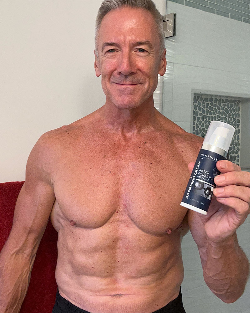 Gray-haired man smiling, holding a bottle of Particle Men's Formula AB Firming Cream.
