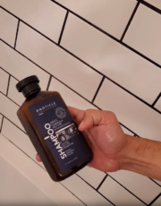 Hand holding a Particle shampoo bottle in front of tiled wall.
