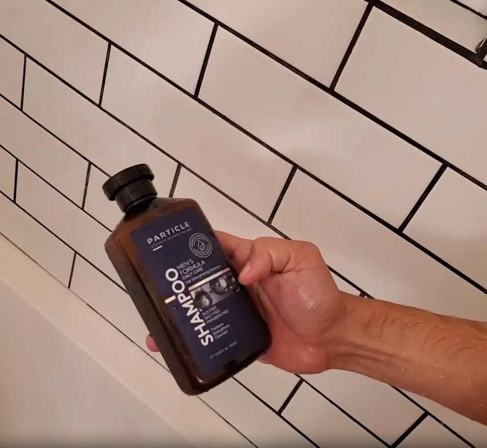 Hand holding a Particle shampoo bottle in front of tiled wall.